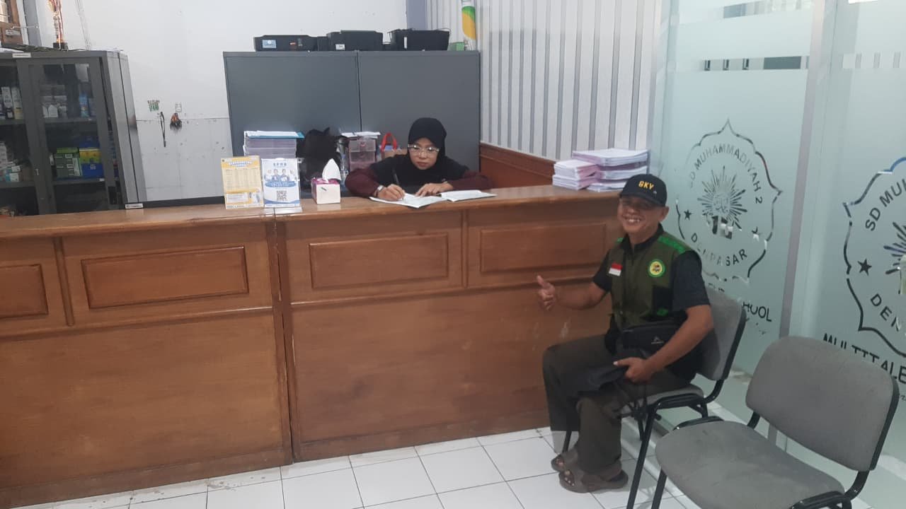 Petugas sekolah mencatat dokumen di meja resepsionis saat seorang relawan yayasan duduk di dekatnya.